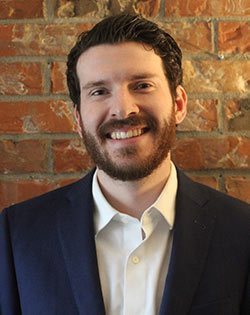 Picture of Gregory: Greg is smiling. He has brown curly hair and a beard. He is wearing a white collared shirt and navy jacket and stands in front of brick wall. 